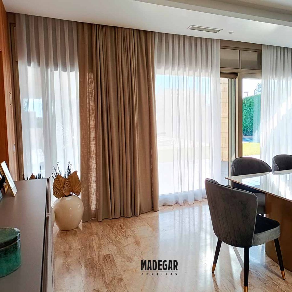 Unas cortinas elegantes en un comedor con grandes ventanales cubiertos por unas cortinas dobles hechas a medida por Cortinastudi, las dobles cortinas están formadas por una tela transparente tipo visillo en lino color natural y una tela más tupida de color tostado con mucha caída, las cortinas en color arena se pueden cerrar totalmente para conseguir intimidad